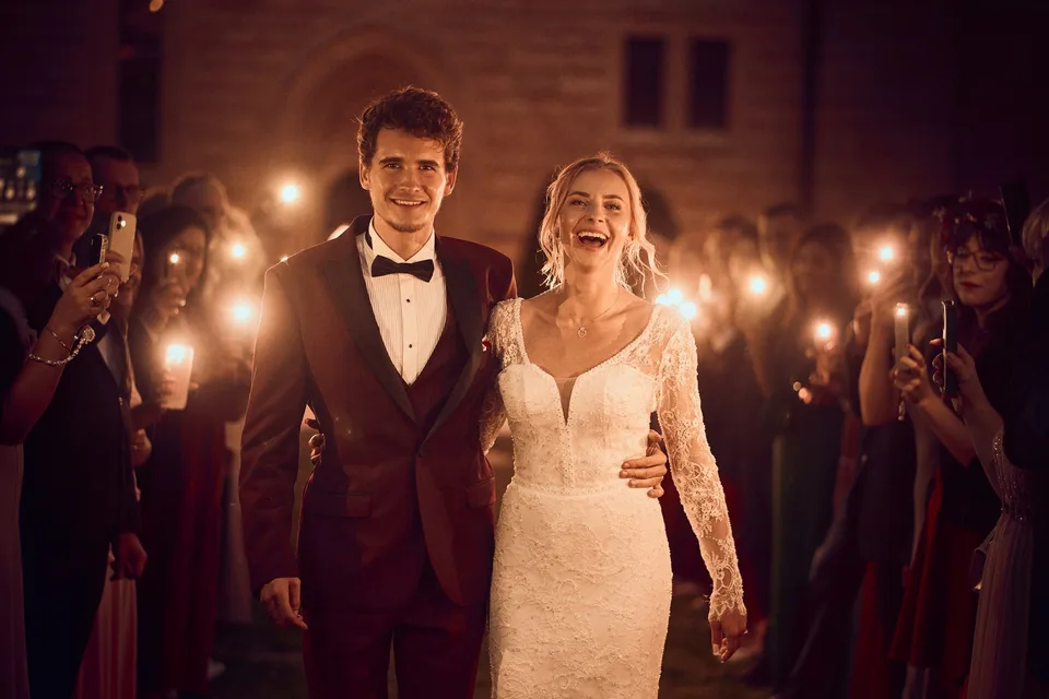 Roześmiana para młoda idzie nocą między gośćmi trzymającymi światła